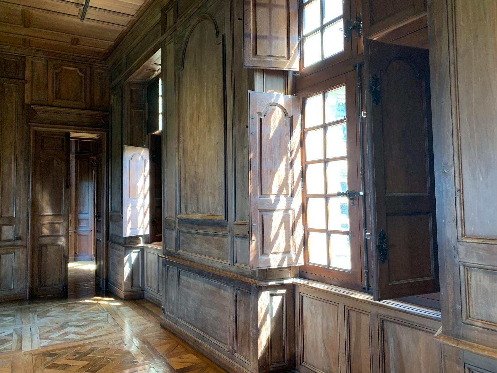 Sunlit wooden interior with ornate windows in a historic French château, featuring classic woodwork and stone elements.