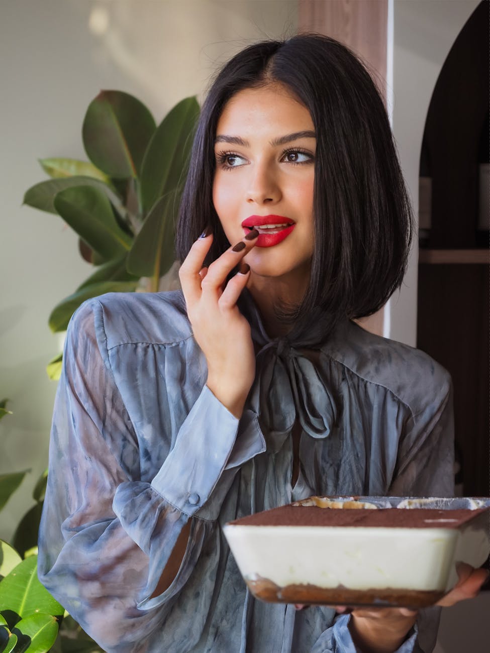 Young woman with red lipstick savoring a delicious dessert indoors with a plant backdrop.