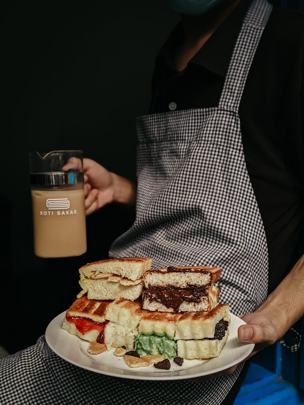 A close-up of a man holding a plate of gourmet roti bakar and a beverage, wearing a checkered apron.