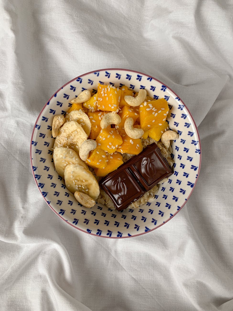 Healthy breakfast bowl with mango, banana, cashews, and chocolate on white fabric.